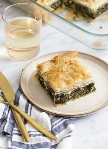 A photo of a piece of spanakiopita on a white marble table with a glass of white wine.