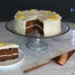 A photo of a carrot and pineapple cake on a cake plate, with a piece removed.