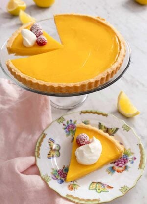 A lemon tart on a cake stand with a slice plated in front.