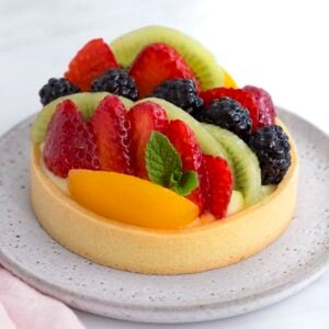 A photo of a classic Fruit tart on a light grey plate with a sprig of mint tucked in.