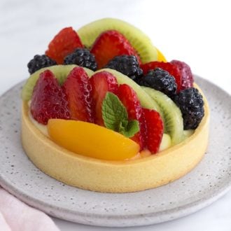 A photo of a classic Fruit tart on a light grey plate with a sprig of mint tucked in.