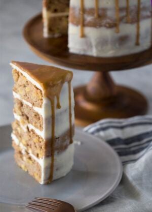 A piece of apple spice cake on a plate with the rest of the cake behind it.