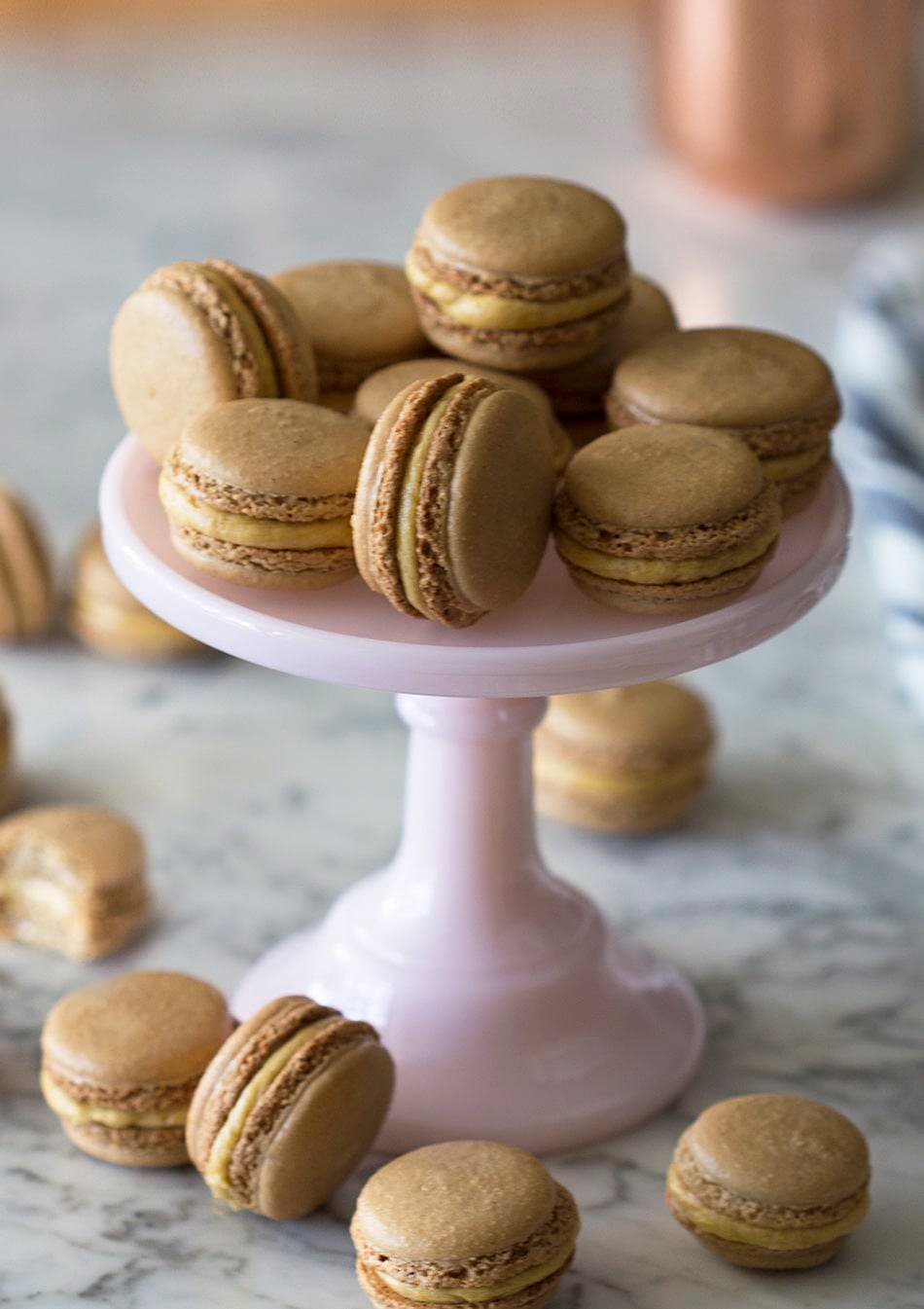 Pumpkin macarons piled onto a cake plate with others surrounding it.