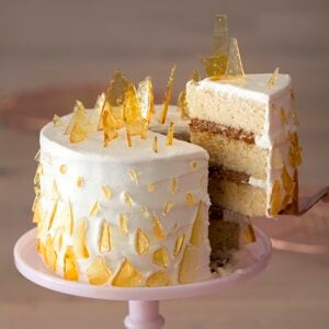 A photo showing a dulce de leche cake on a pink cake stand with a piece being removed. The cake is covered in sugar shards