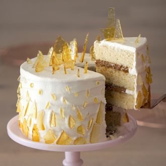 A photo showing a dulce de leche cake on a pink cake stand with a piece being removed. The cake is covered in sugar shards