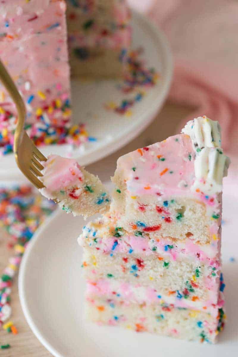 Photo of a piece of pink funfetti cake on a white plate being eaten.