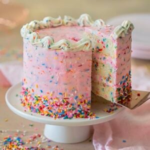 A pink birthday cake covered in rainbow sprinkles on a white cake stand with a piece being removed