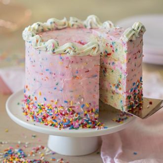 A pink birthday cake covered in rainbow sprinkles on a white cake stand with a piece being removed