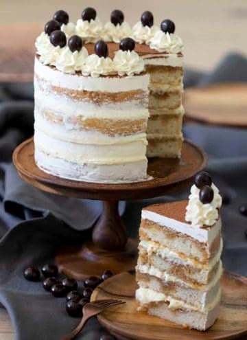 A tiramiisu cake on a wooden cake stand with a piece in the foreground.
