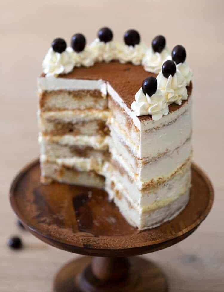 A tiramisu cake topped with cocoa powder on a wooden cake stand.