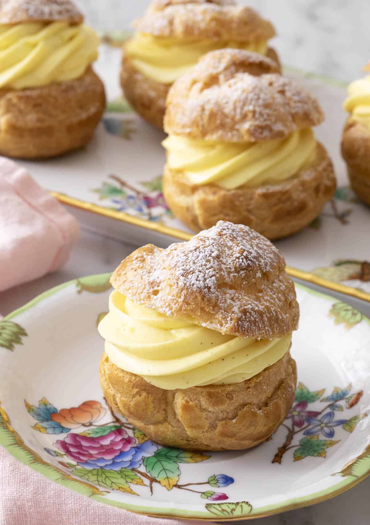 A cream puff with a dusting of powdered sugar on a plate in front of a platter.