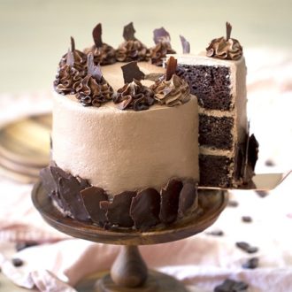 A photo of a chocolate cake on a wooden cake stand with a piece being removed.