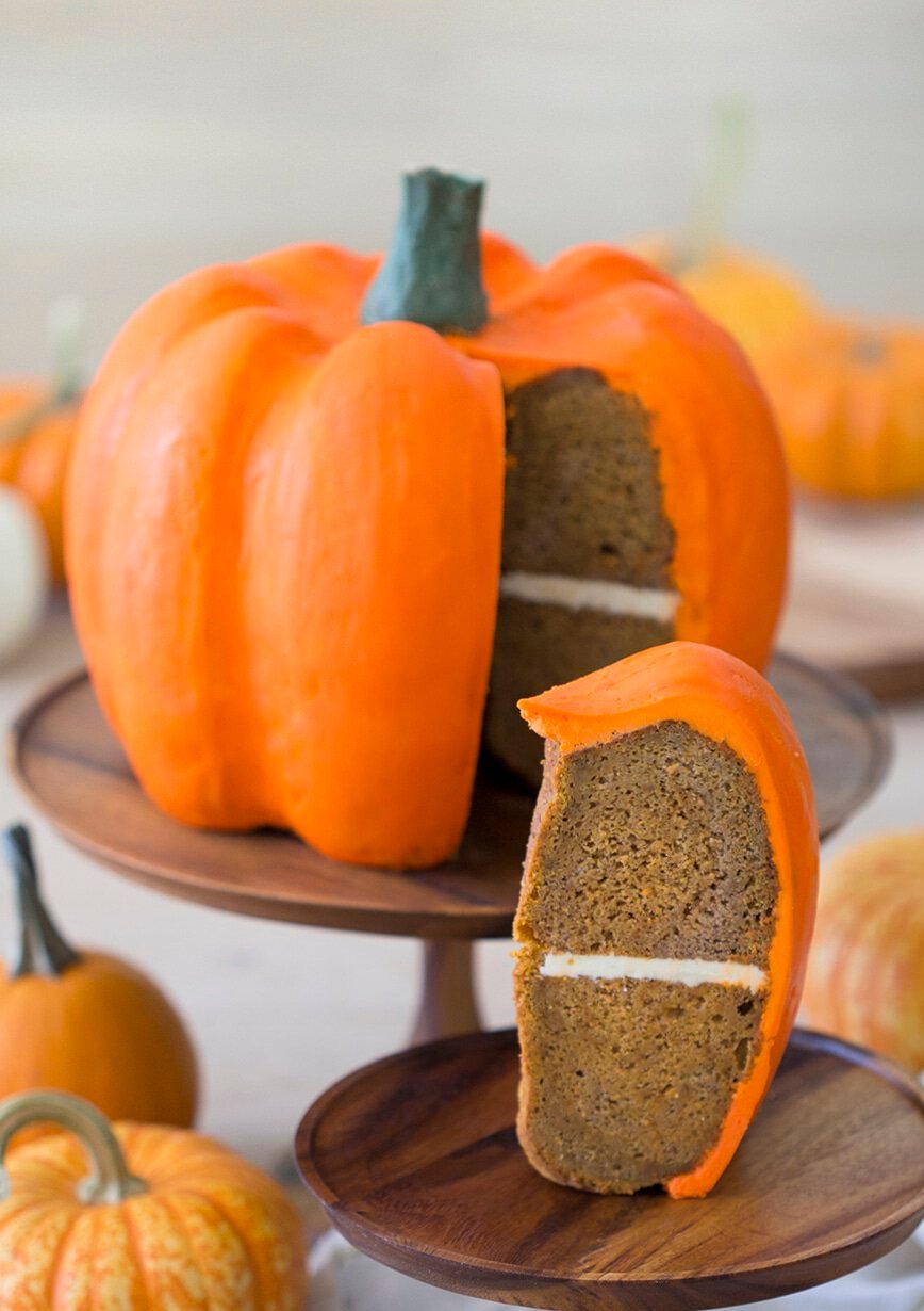 Photo of a large orange pumpkin cake with a piece in the foreground.