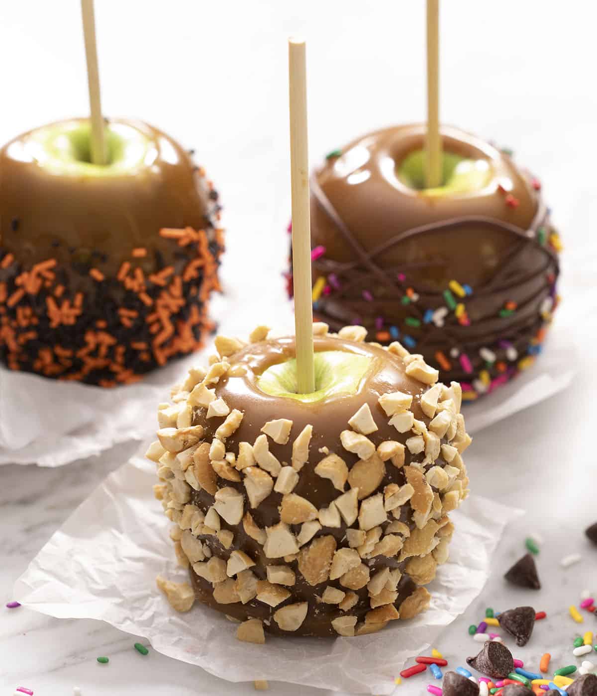 Three caramel apples decorated with chocolate nuts and sprinkles.