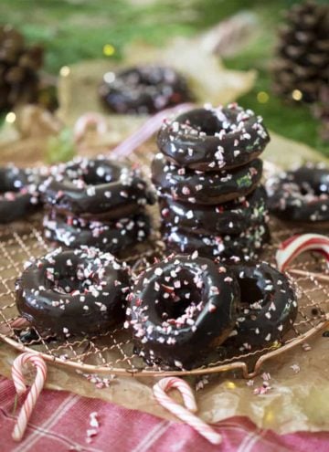 A photo of chocolate peppermint donuts, in a pile on a wire rack, with candy canes around them.
