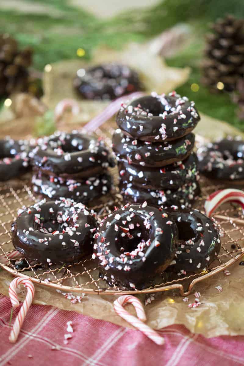 A photo of chocolate peppermint donuts, in a pile on a wire rack, with candy canes around them.