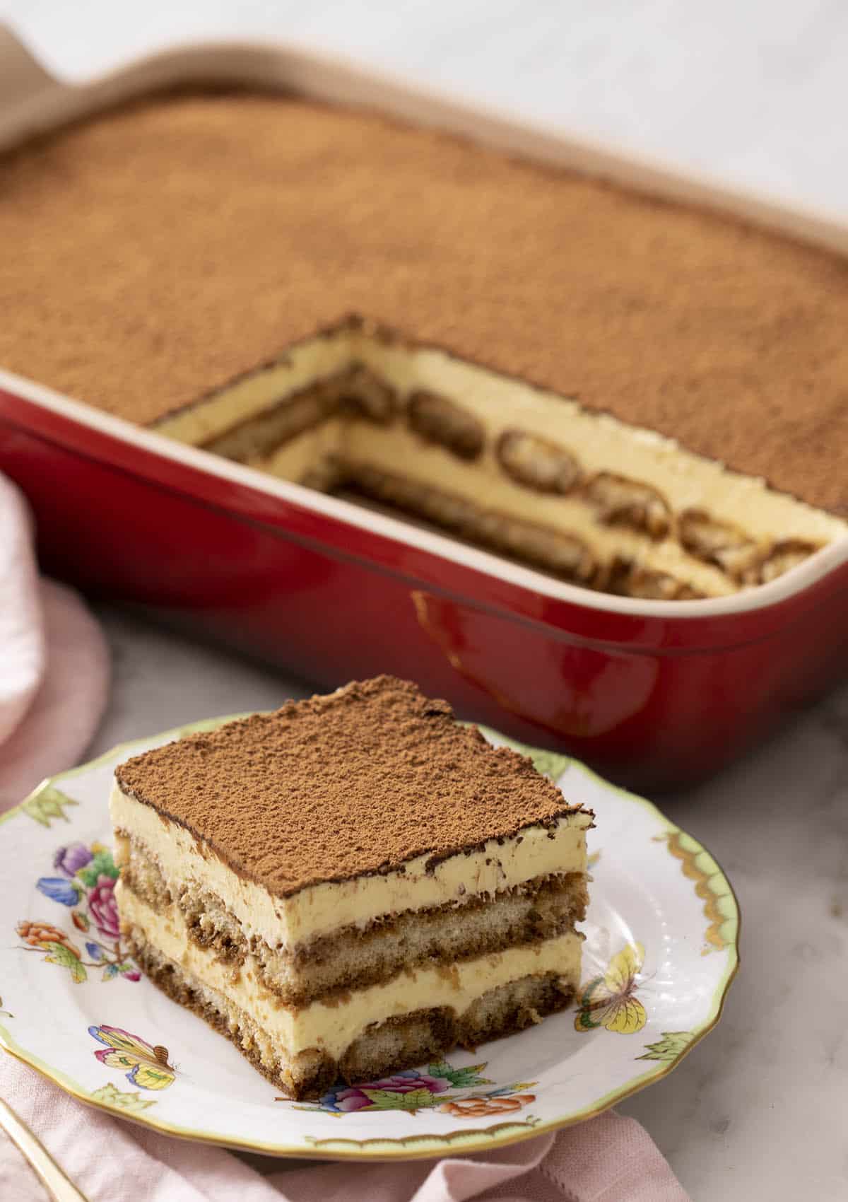 A piece of tiramisu in front of a red serving dish.