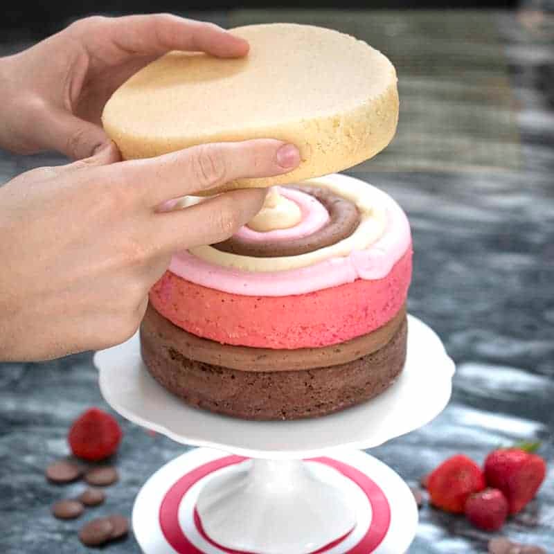 Photo of a Neapolitan cake being assembled on a white cake stand.