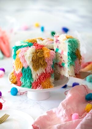 Photo of a colorful shag cake on a white cake stand with a piece being removed