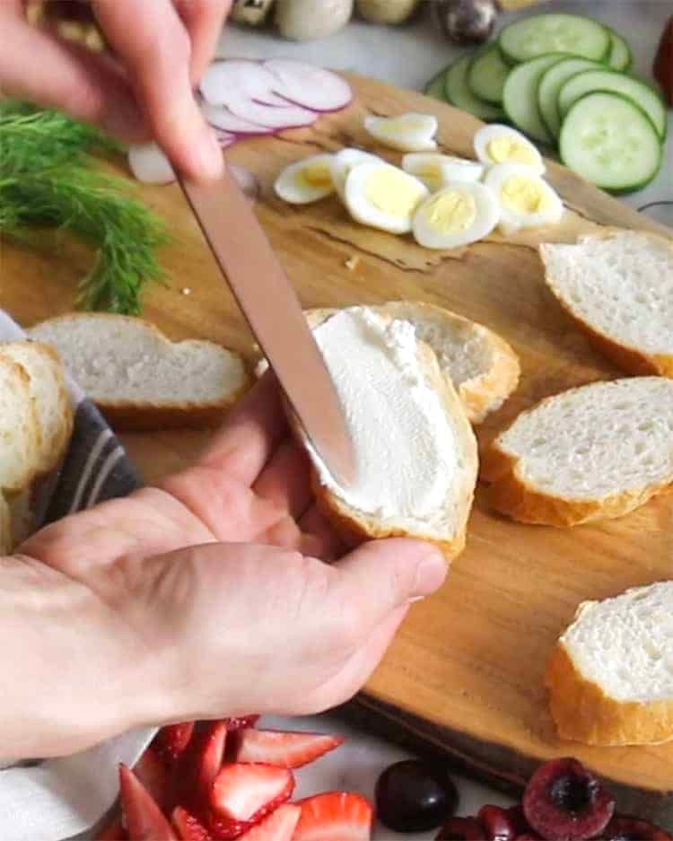 Cheese being spread onto a slice of French bread with a knife.