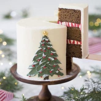 A photo of a gingerbread cake with a painted buttercream pine tree on the front and a piece being removed.