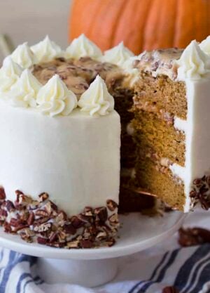 A slice of pumpkin pecan cake being lifted from the rest of the cake from a white cake stand.