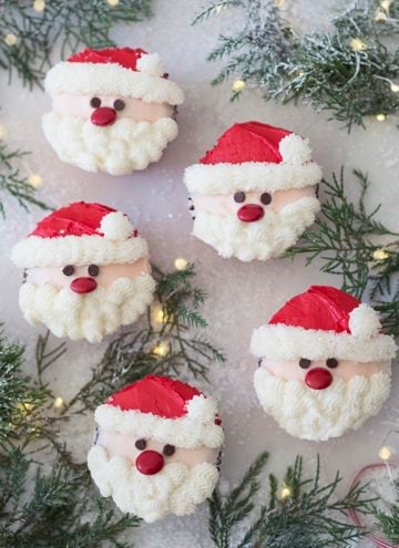 A photo showing a group of santa cupcakes on a white marble table with Christmas lights and evergreens