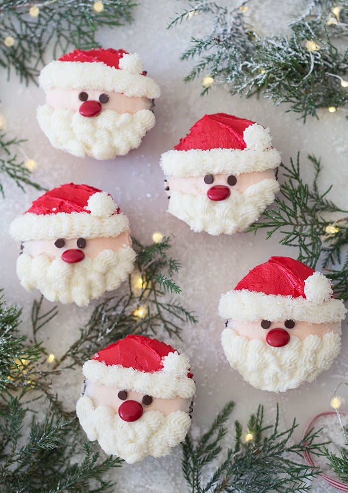 A photo showing a group of santa cupcakes on a white marble table with Christmas lights and evergreens