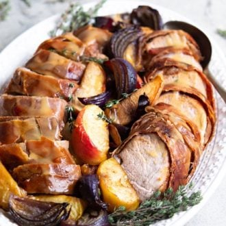 A photo of bacon-wrapped pork tenderloin slices arranged on a platter with apples potatoes and onions.