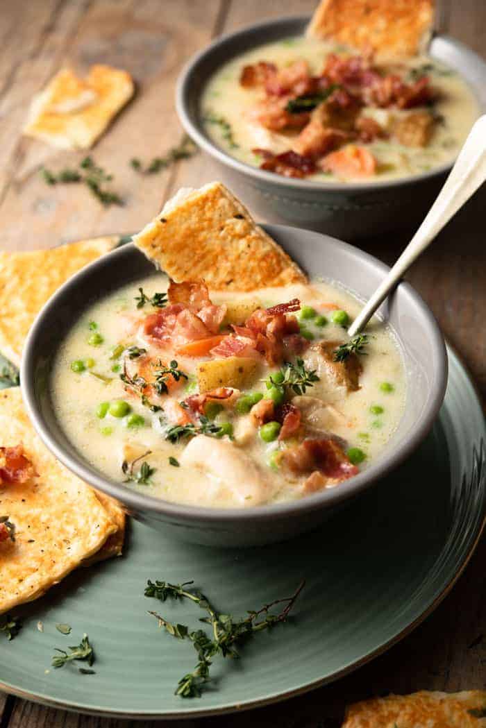 A close up of chicken pot pie soup in a gray bowl topped with thyme, bacon and crackers