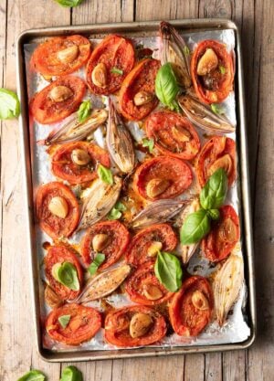 Overhead view of roasted tomatoes with shallots, garlic, and basil on a sheet pan.