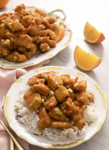 Rice and orange chicken on a plate with orange wedges in the background
