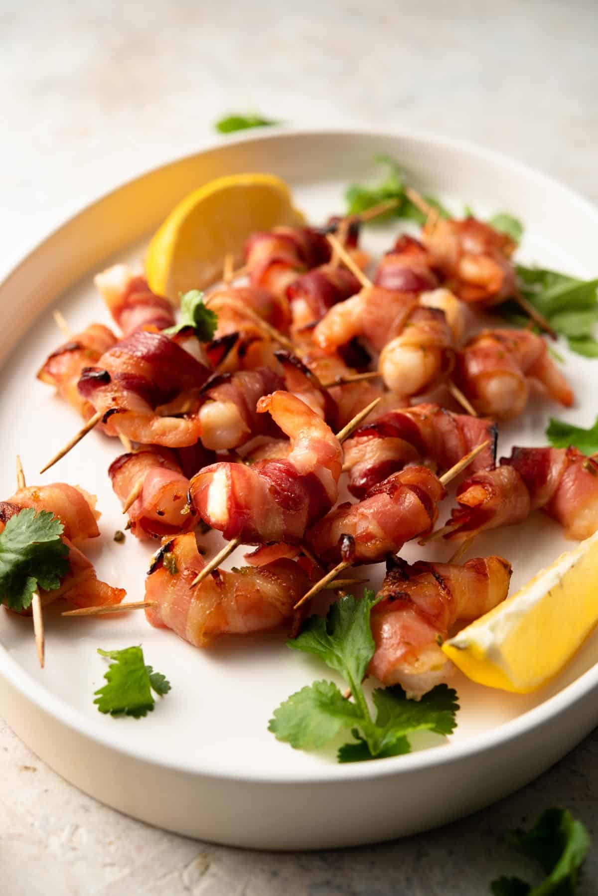 A side shot of bacon wrapped shrimp on a white plate with lemon wedges