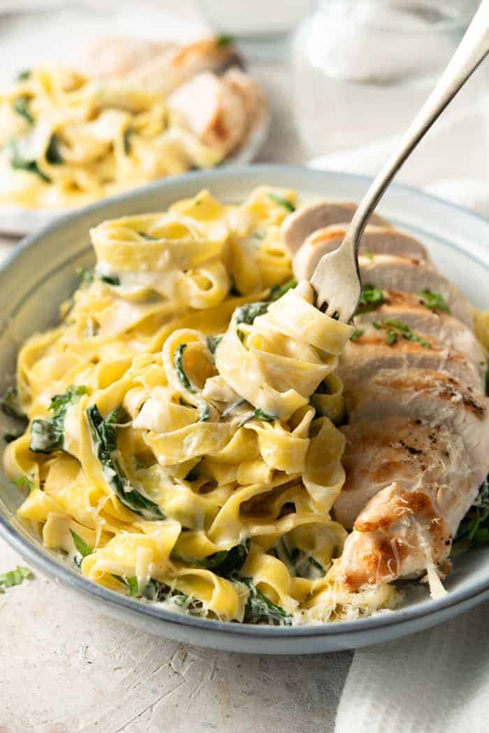 A close up of a fork picking up some chicken alfredo from a bowl