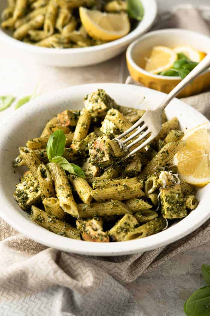 A close up of pesto chicken pasta in a white bowl with a fork and lemon wedges