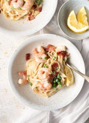 An overhead shot of shrimp alfredo in a white bowl with bacon and peas