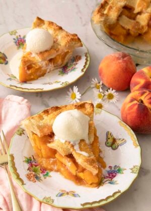 Two pieces of peach pie on porcelain plates with ice cream on top.