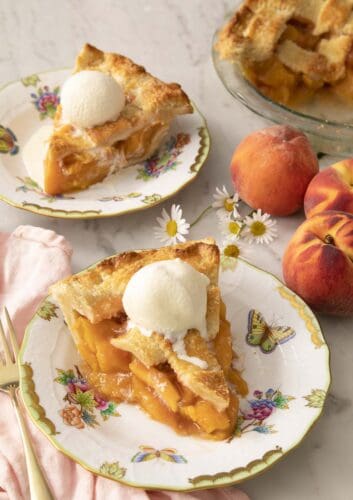 Two pieces of peach pie on porcelain plates with ice cream on top.