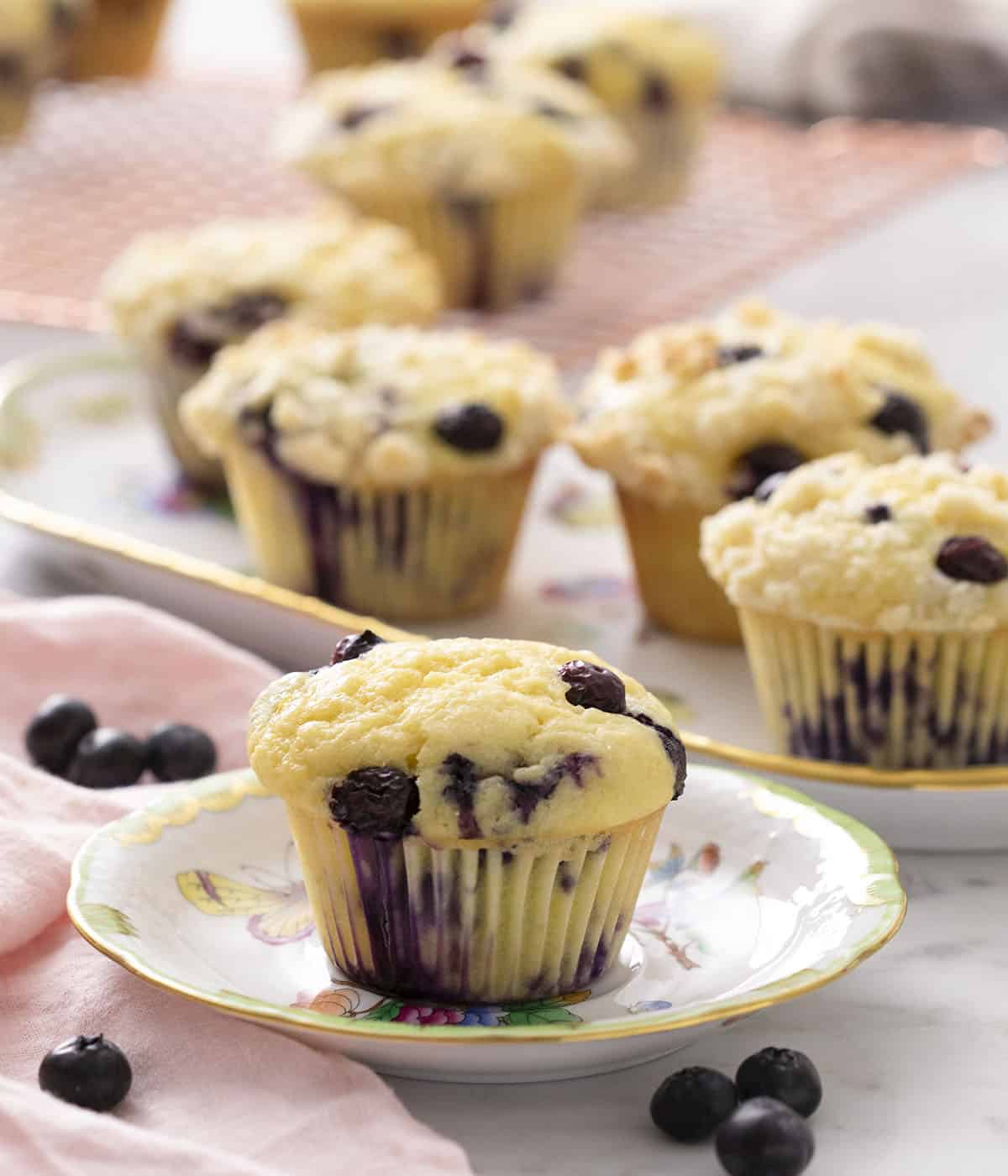 A blueberry muffin on a small plate with a platter of more muffins in the background.