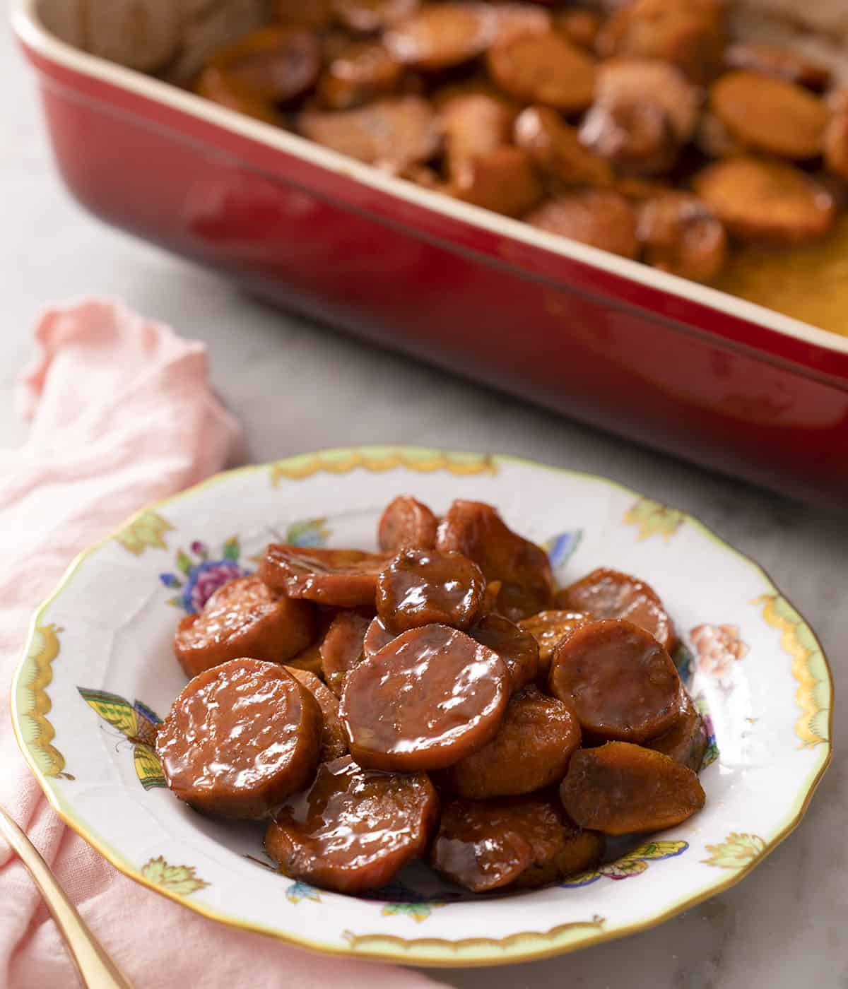 A porcelain plate with candied yams in front of a red baking dish.