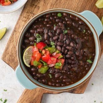 An overhead view of a bowl of Instant Pot black beans topped with some salsa, lime, and herb garnish on top of a wooden serving board.