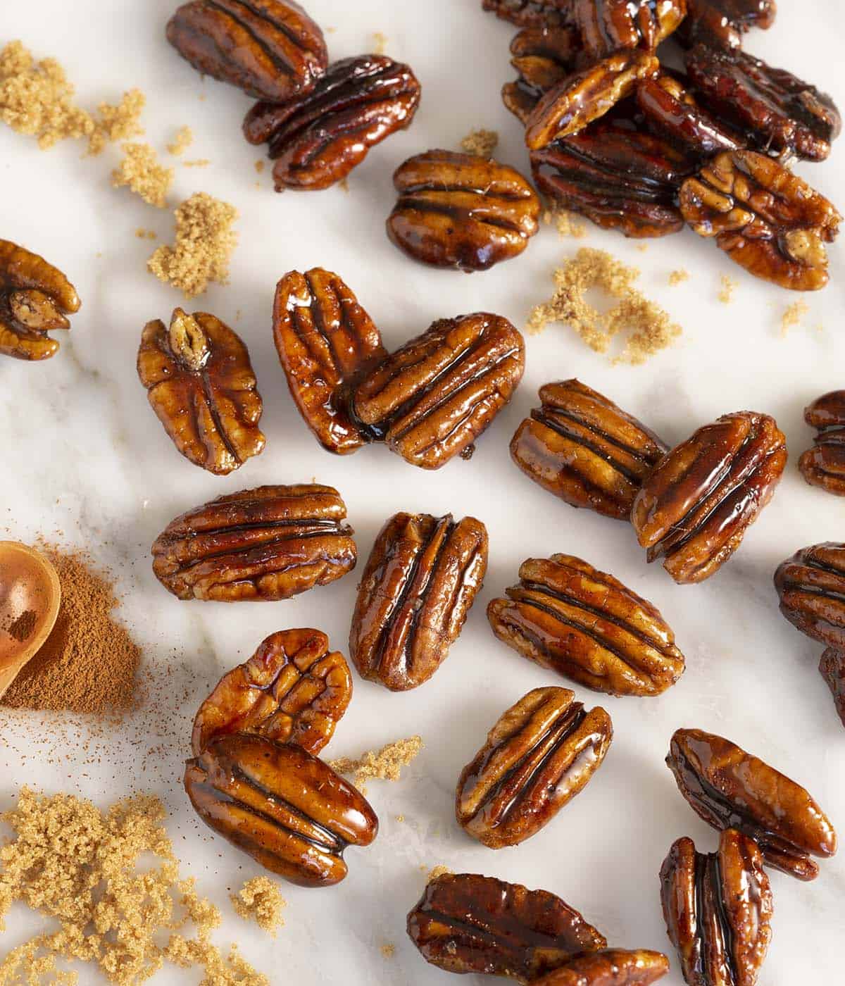 A group of candied pecans on a white marble table with cinnamon and brown sugar in frame.