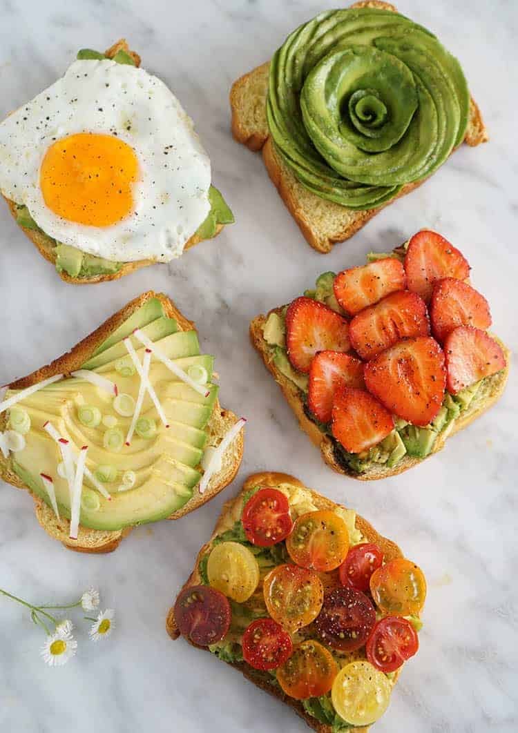 Five pieces of avocado toast on a white marble surface, each with different toppings
