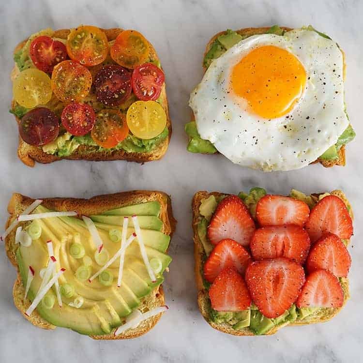 Four pieces of avocado toast on a white marble table, each piece is topped differently