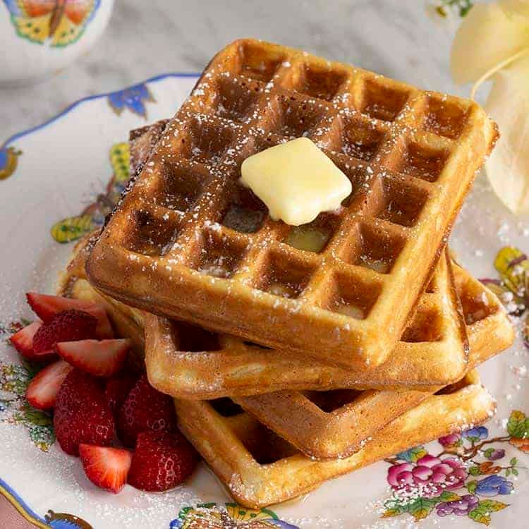 Four Waffles stacked on a plate with a pat of butter on top and strawberries as the base
