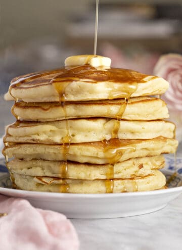 A tall stack of pancakes topped with a pat of butter with syrup being poured on top.