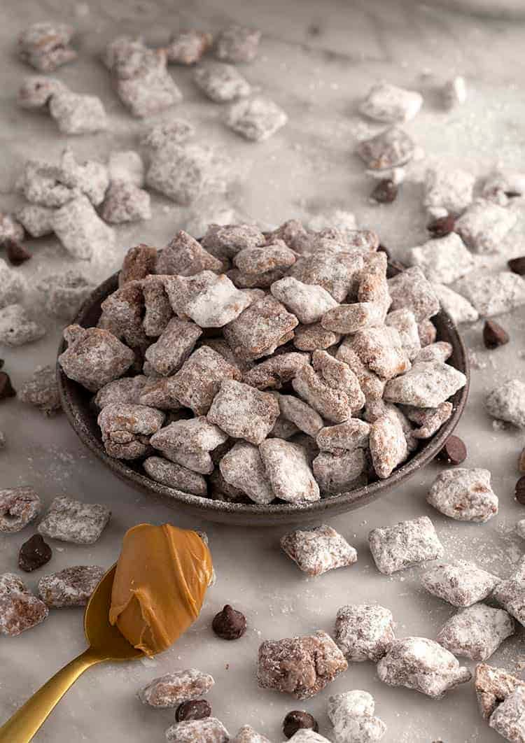 A bowl of puppy chow on a white marble table with a spoon of peanut butter.