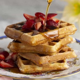 A stack of waffles with cut up strawberries getting maple syrup poured on it.