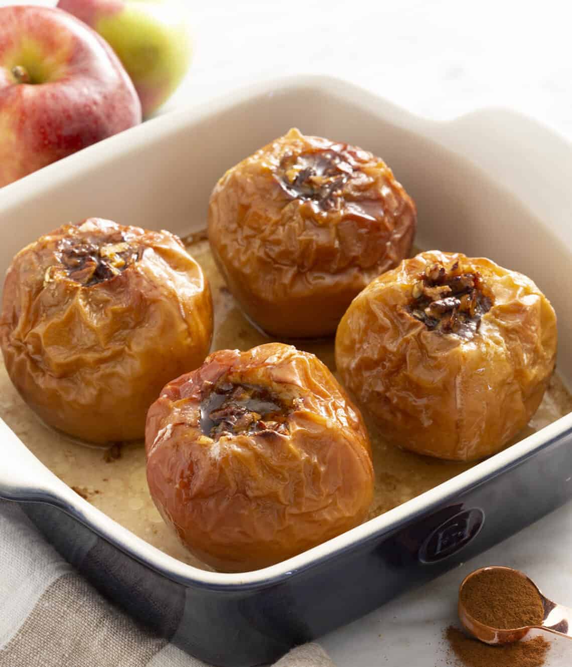 A square baking with four baked apples. A spoon of cinnamon in front and two apples in the back.