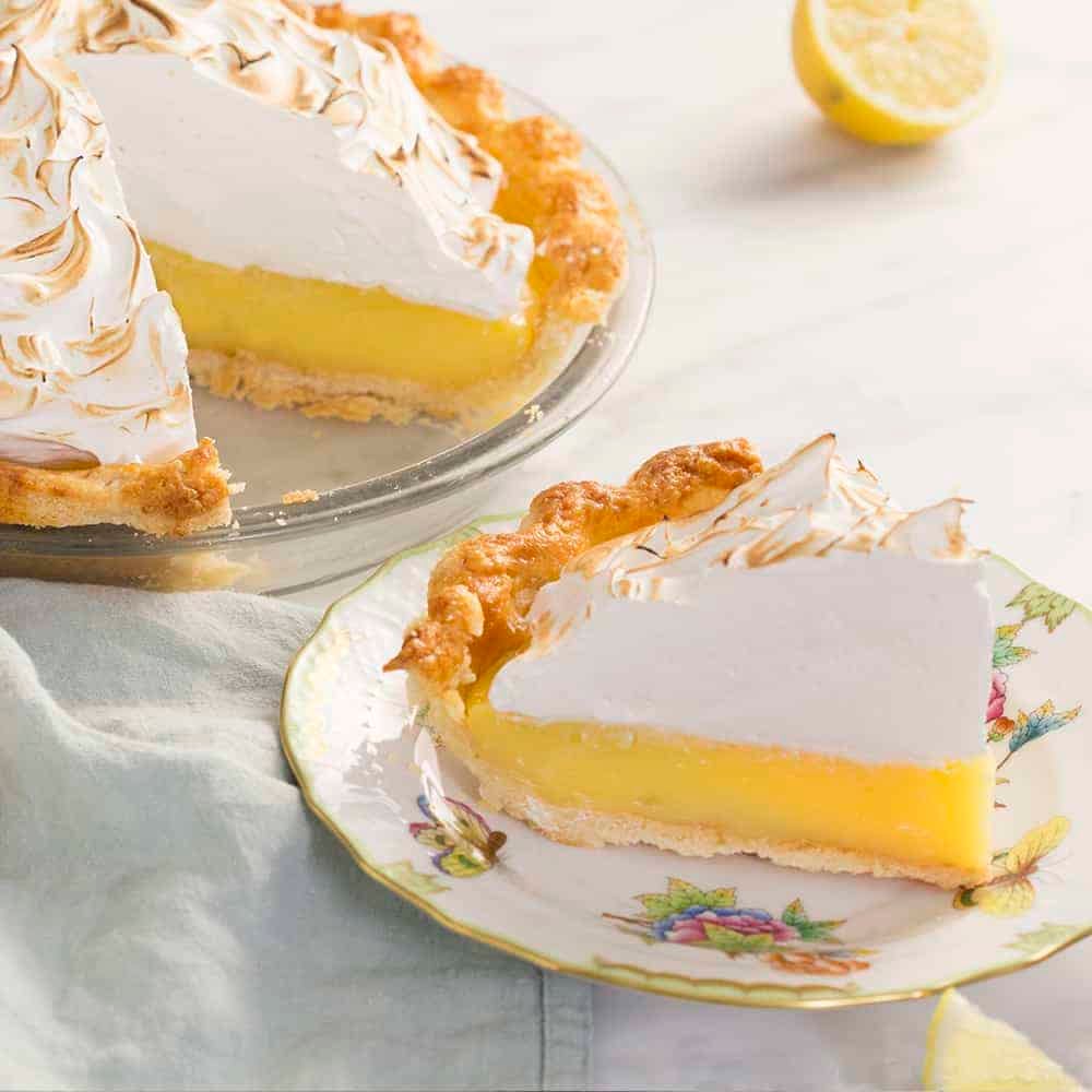 A lemon mering pie ppiece next to the cut pie on a marble table.